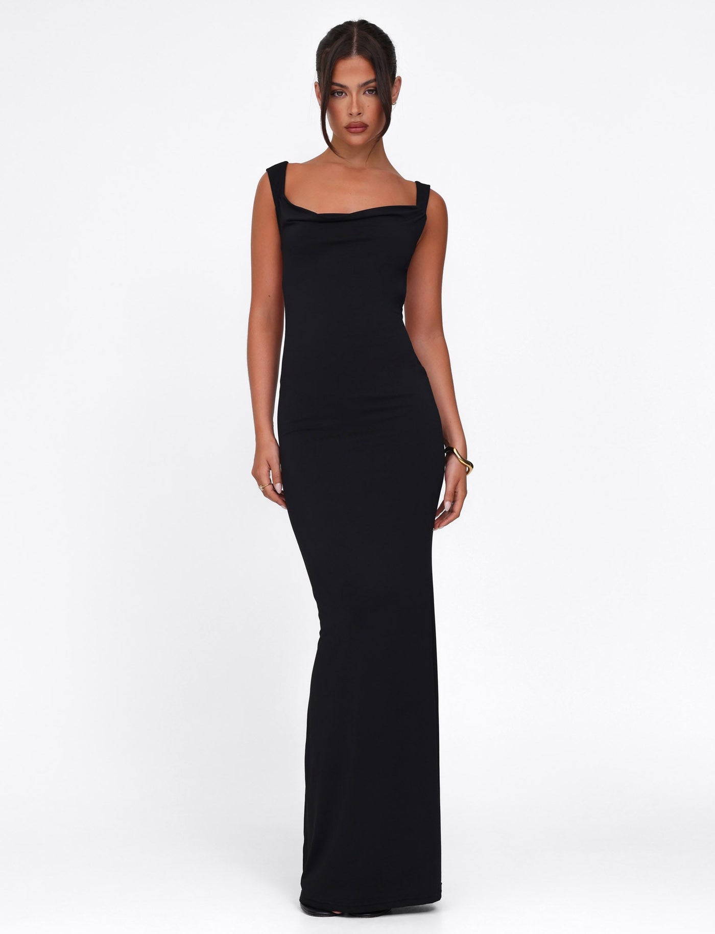 A woman stands against a white background wearing the sleek MORANA DRESS - BLACK by TIGERMIST, a long bodycon maxi dress with wide straps. Her hair is pulled back, and she displays a neutral expression.