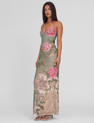 A woman models the TIGERMIST JULIANNA DRESS in Green Rococco Floral with large pink flowers, a long fitted maxi dress with thin straps. She pairs it with white open-toe heels, her long brown hair styled straight, against a plain background.