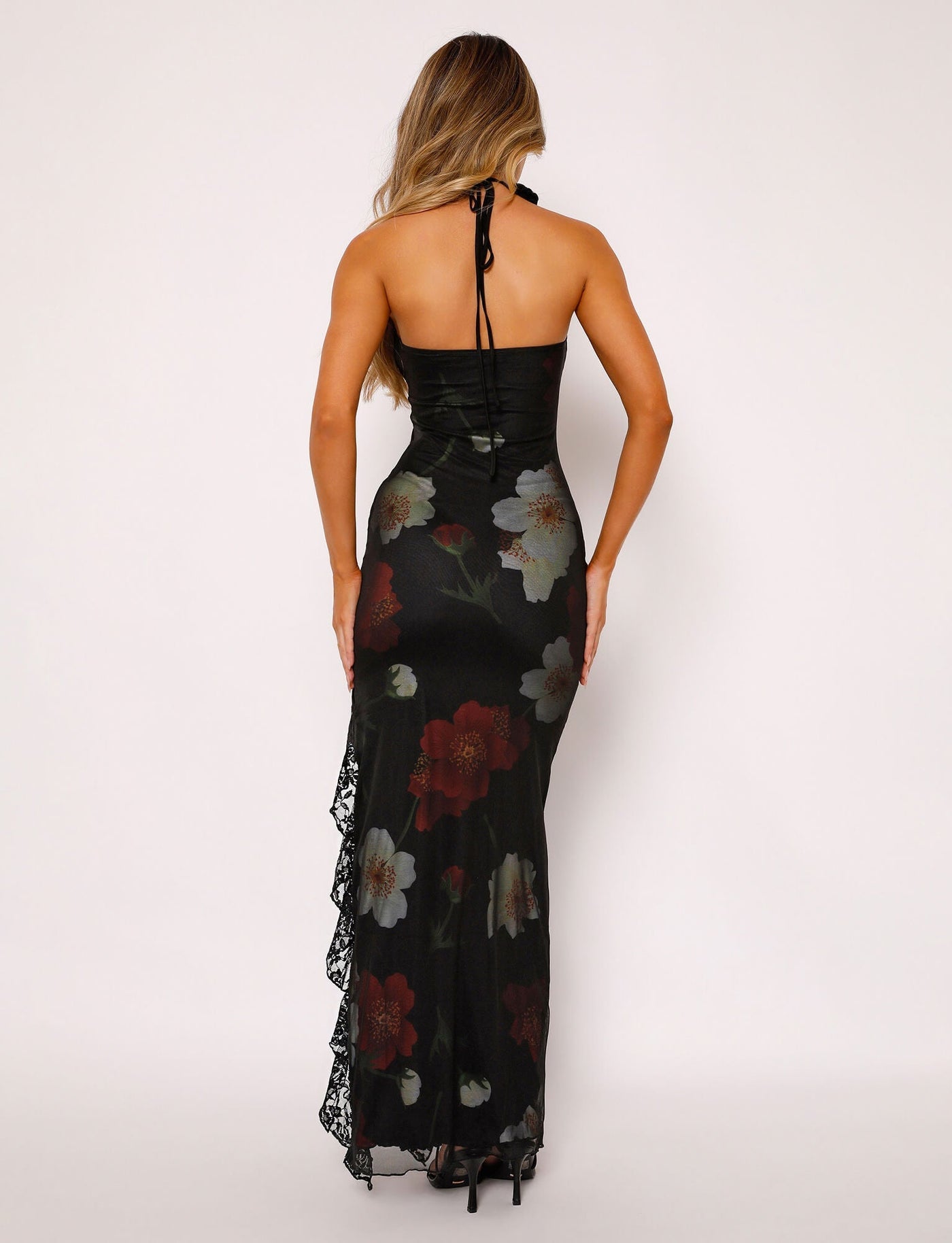 A woman with long hair is seen from the back, wearing the TIGERMIST THEO MAXI DRESS - RED : MACRO FLORAL. The black halter-neck dress is adorned with large red, white, and green floral patterns and features lace detailing on the left side. She complements her outfit with black high heels against a plain white background.