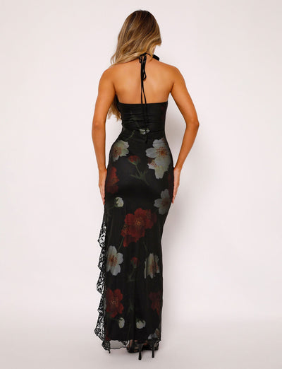 A woman with long hair is seen from the back, wearing the TIGERMIST THEO MAXI DRESS - RED : MACRO FLORAL. The black halter-neck dress is adorned with large red, white, and green floral patterns and features lace detailing on the left side. She complements her outfit with black high heels against a plain white background.