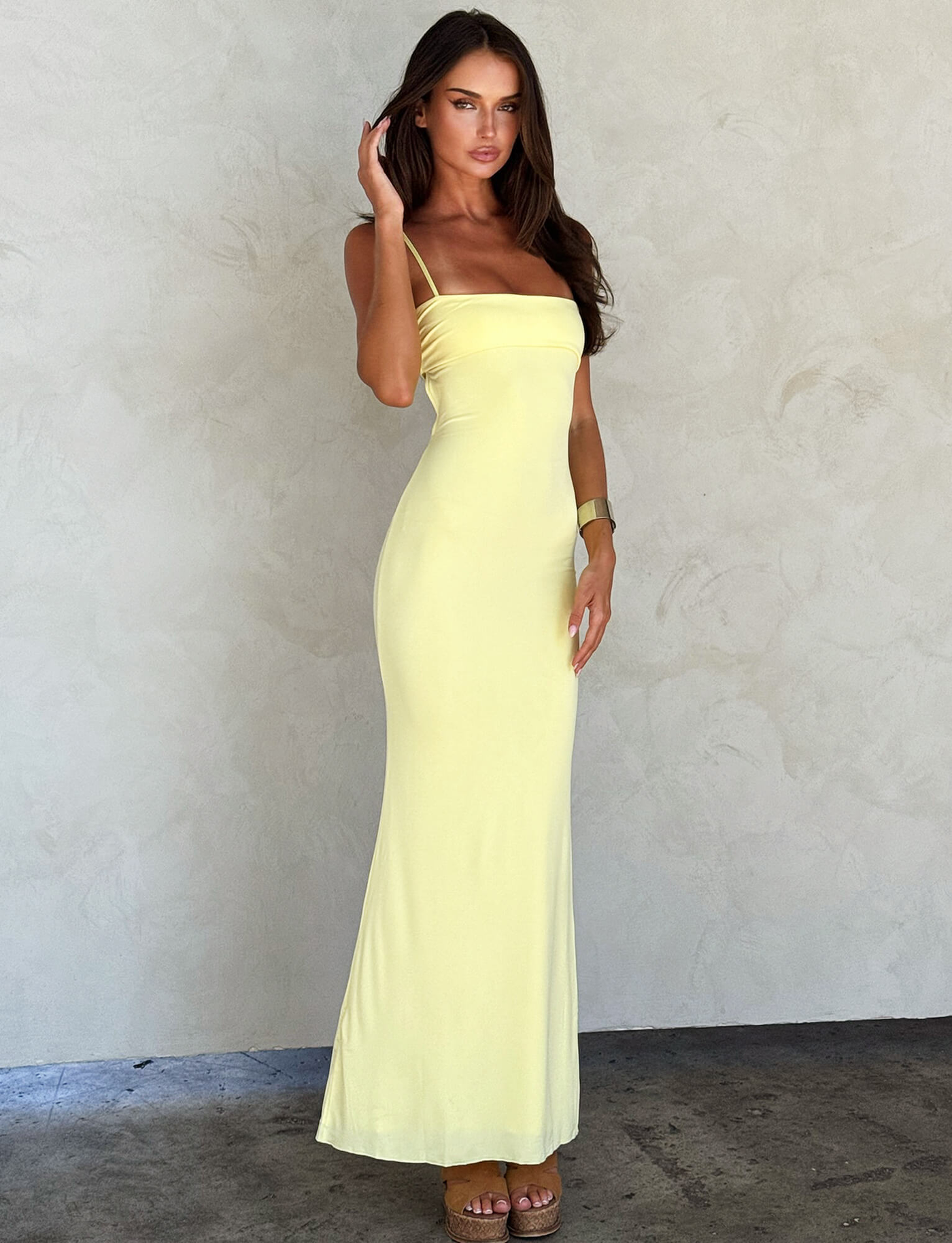 Against a light textured wall, a woman in the TIGERMIST CARA DRESS - YELLOW : PASTEL CITRUS with thin straps and wedge sandals channels boho vibes. Her long brown hair frames her face as she raises one hand near it.