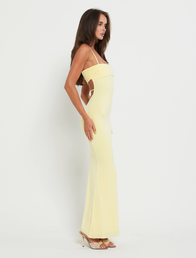 A woman stands sideways against a white background, wearing the TIGERMIST CARA DRESS in Pastel Citrus— a pale yellow, floor-length, sleeveless dress with thin straps and open sides—paired with beige high-heeled shoes.