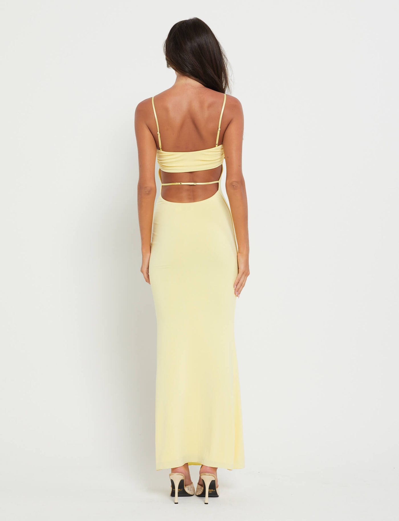 A woman faces away in the TIGERMIST CARA DRESS - YELLOW : PASTEL CITRUS, a long, strappy dress with an open back and horizontal cutout. Her high heels and flowing dark hair create a subtle bohemian vibe against a plain white background.