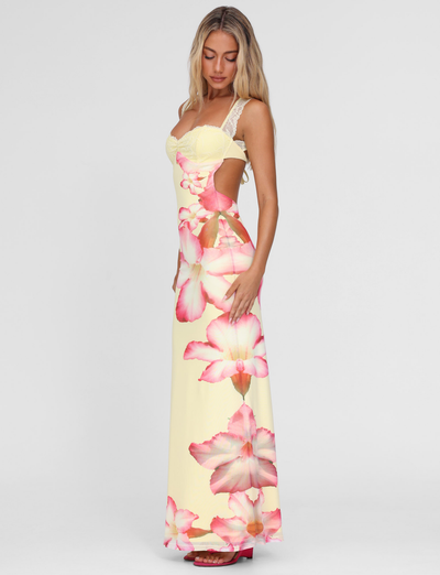 A woman with long blonde hair models the TIGERMIST NYOMI MAXI DRESS in Yellow Hibiscus Macro Floral—a sleeveless, fitted dress with lace straps and open back—against a plain white background.