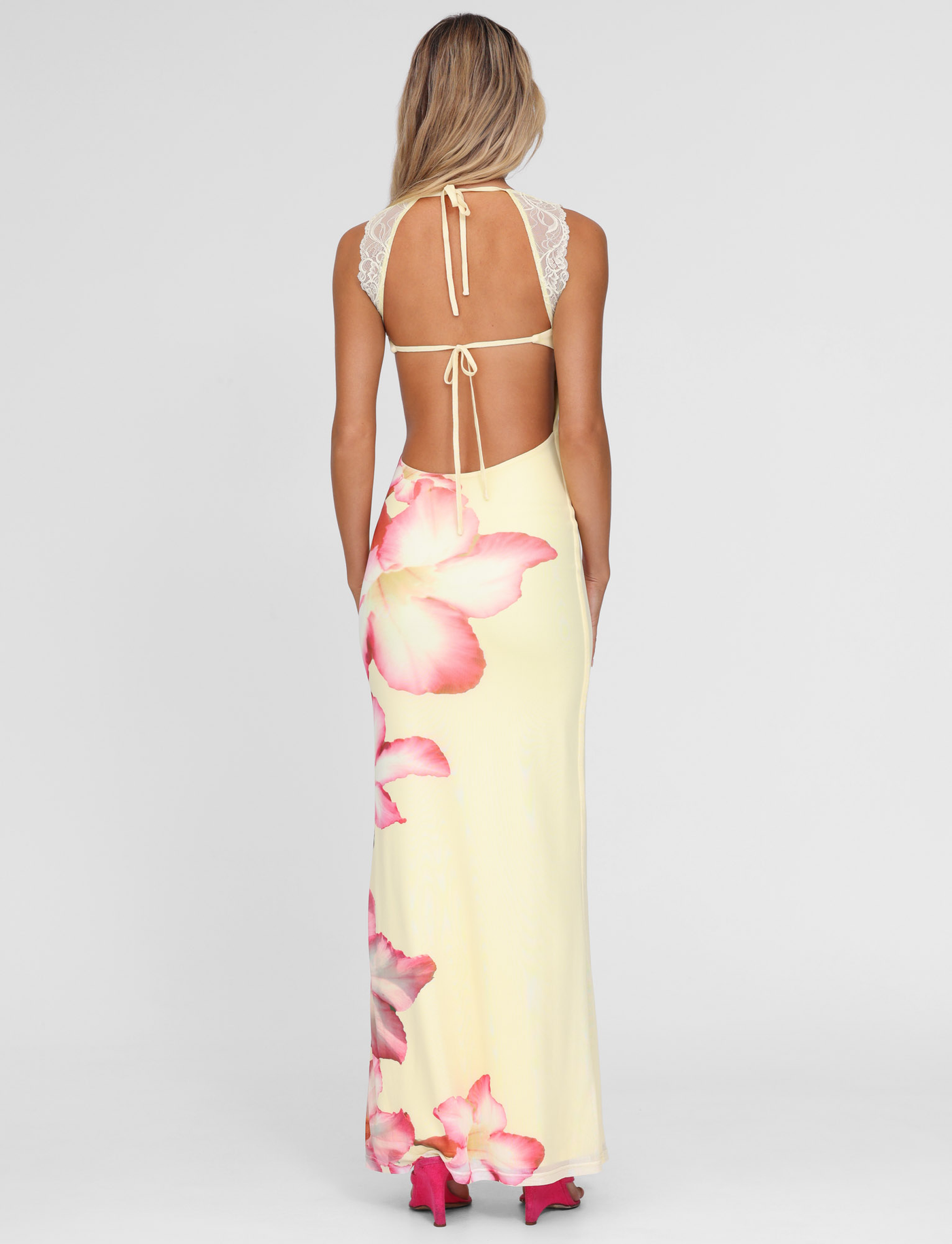 A woman with long blonde hair wears the TIGERMIST NYOMI MAXI DRESS in yellow hibiscus macro floral, featuring a backless design and lace straps, paired with pink heels against a plain light background.